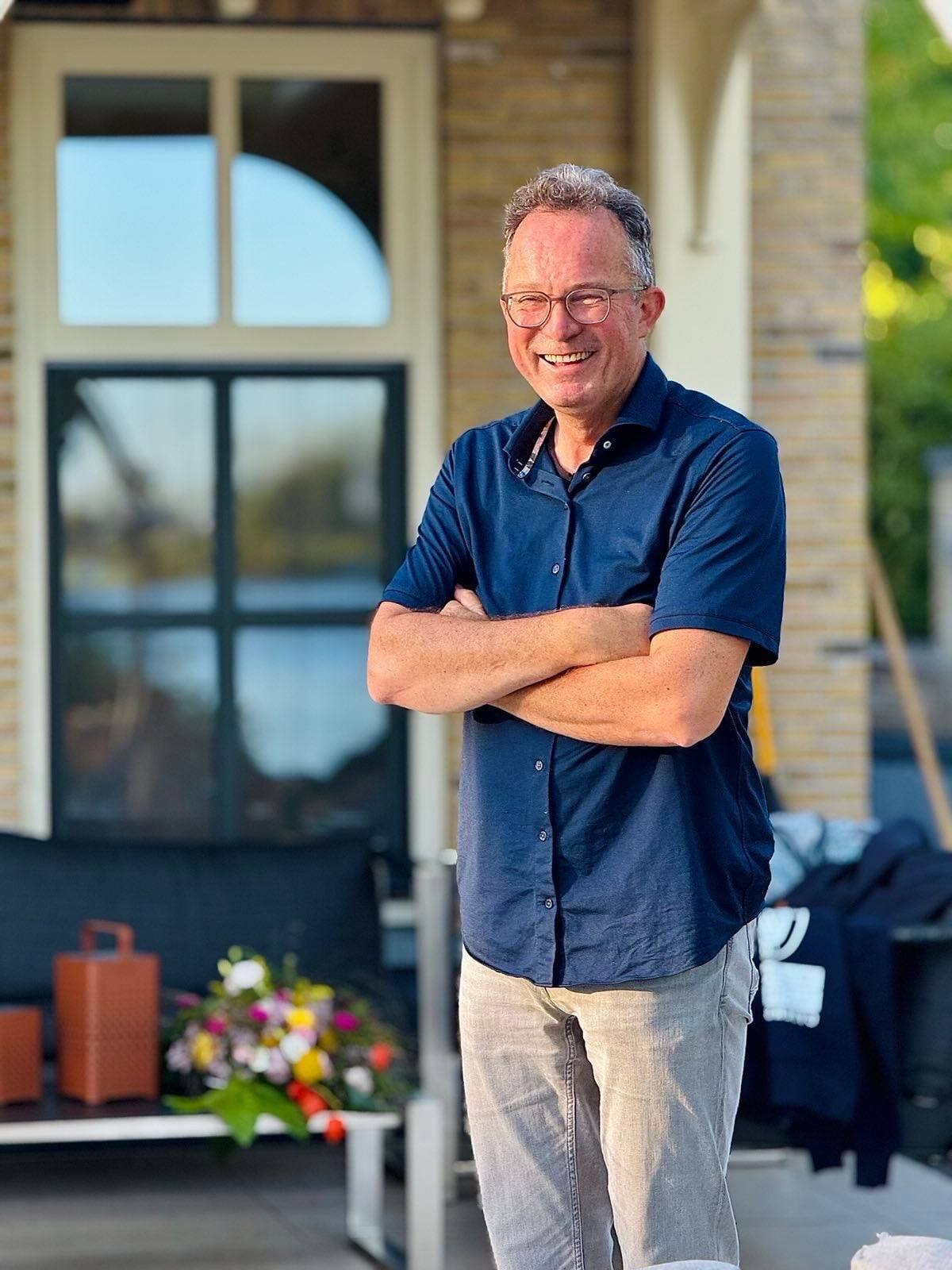Een man in een donker overhemd, met de armen over elkaar, lachend, voor een bakstenen huis in de avondzon.