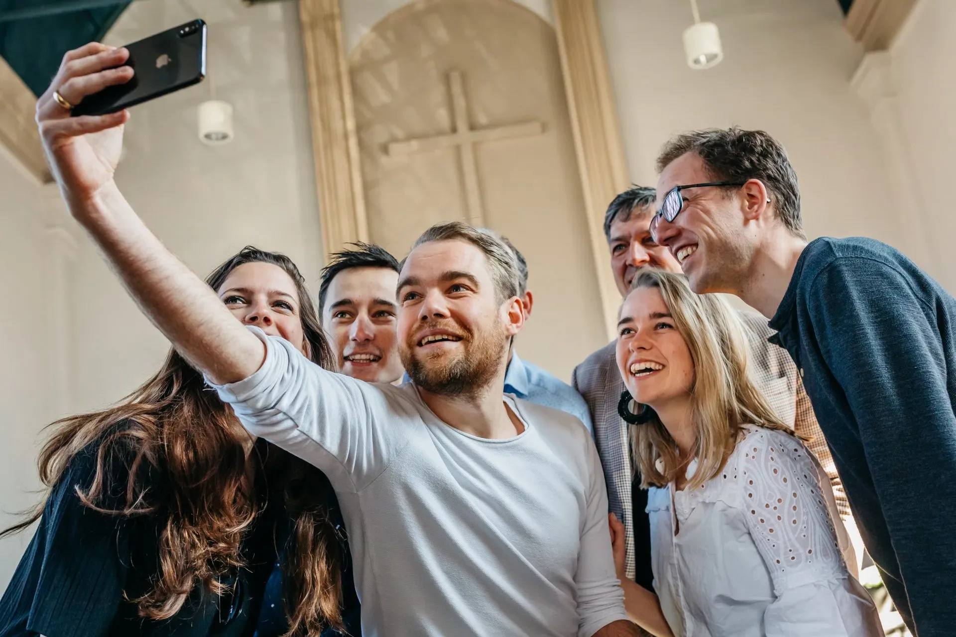 A group of people crowded together for a selfie, laughing, in a tall, light church interior.